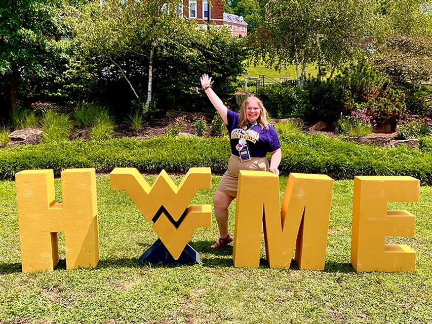 Raeanne Beckner stands in front of a huge Home sign with the flying WV