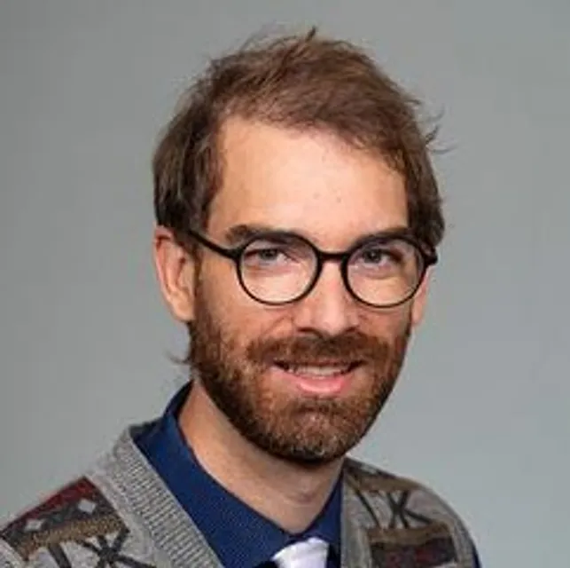 Male with a full beard, thick plastic rimmed glasses is posing for a studio shot while wearing a patterned sweater vest, button down shirt and a tie.