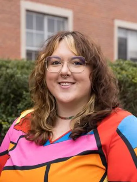 Woman with wavy shoulder length hair wearing octagon shaped wire rimmed glasses and a bold color block shirt smiles for the camera.