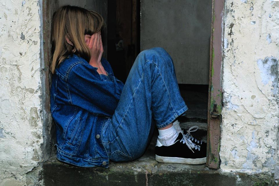 A preteen girl wearing jeans and a jean jacket sits between two walls, burying her face in her hands. 