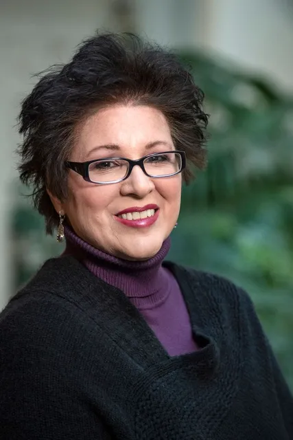 Smiling woman with short brunette hair, dark glasses wears a purple shirt with a black suit jacket. 