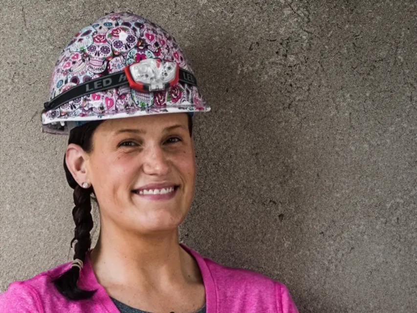 Smiling female with dark eyes and braided, dark brown hair wears a brightly patterned construction helmet. 
